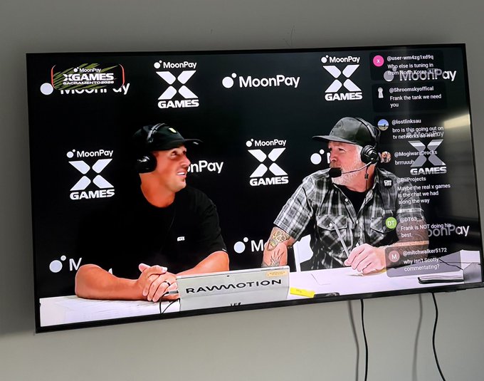 A TV screen displays the MoonPay x X Games live broadcast, featuring two commentators wearing headsets at a desk with "RAW MOTION" visible, while a live chat on the right side shows viewers discussing the real vs. chat conversation and network streaming—aligning with the post about watching MoonPay x Games live.