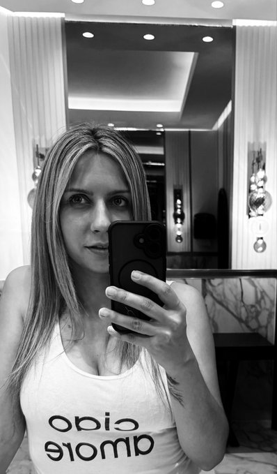 A black-and-white mirror selfie of a woman with long hair wearing a tank top with the word 'Calvin Klein' printed in reverse.