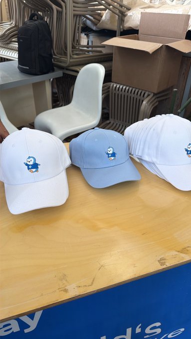 Three baseball caps (white, light blue, and white) featuring a cartoon character logo displayed on a wooden table, with stacked chairs and a cardboard box in the background.