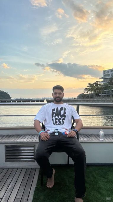 A man sitting on a chair outdoors on a deck by water, wearing a white t-shirt with the text 'FACE LESS' at sunset.