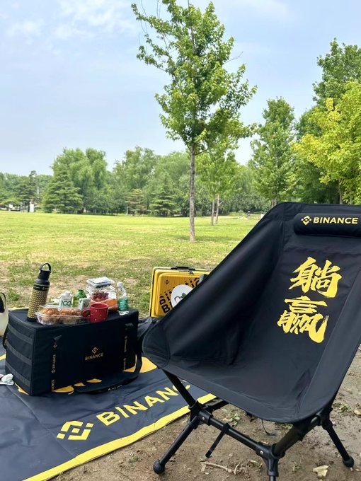 A Binance-branded folding chair and storage box sit on a "BINANCE" mat in a sunny park, surrounded by snacks and drinks for a picnic. The branded gear symbolizes the effortless crypto gains celebrated as $BNB surpassed $700 and outpaced the market cap of Starbucks in June 2024.