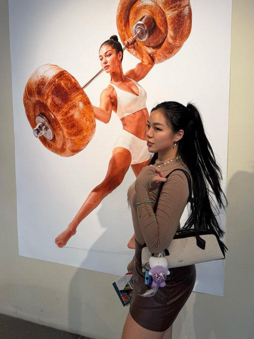 A young woman with a ponytail poses next to a large artwork depicting a muscular figure lifting a barbell made of bagels.