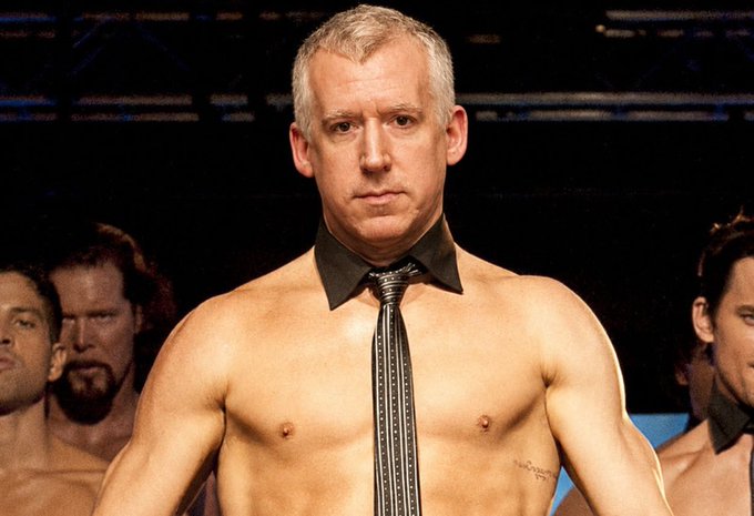 A shirtless, muscular older man with short gray hair wears a black dress shirt collar and a striped necktie, standing in front of other similarly dressed men in a performance or wrestling-style setting.