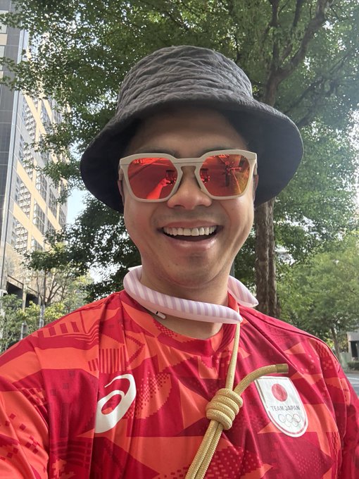A smiling man wearing a gray bucket hat, red-tinted sunglasses, and a red Team Japan Olympic jersey with the Asics logo and Olympic rings.