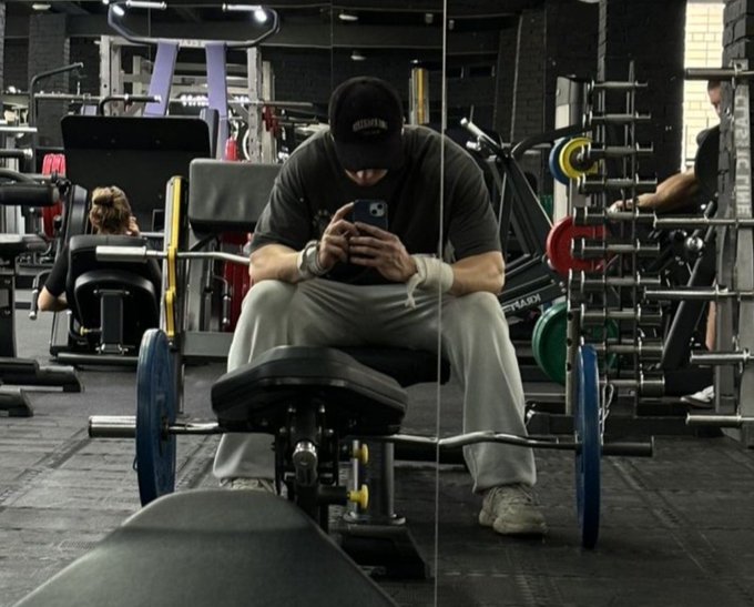 A man in a black cap and gray shirt sitting on gym equipment, looking down at his phone with a barbell in the foreground.