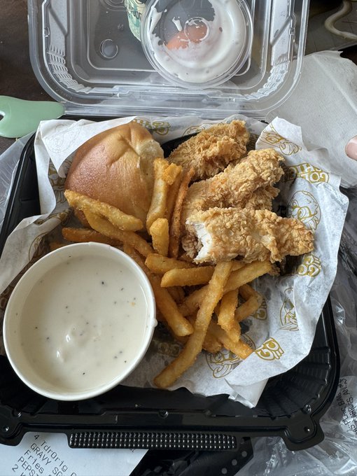 A takeout meal in a black plastic container includes fried chicken tenders, french fries, a dinner roll, and a cup of white dipping sauce, with a receipt and another sauce container visible nearby.