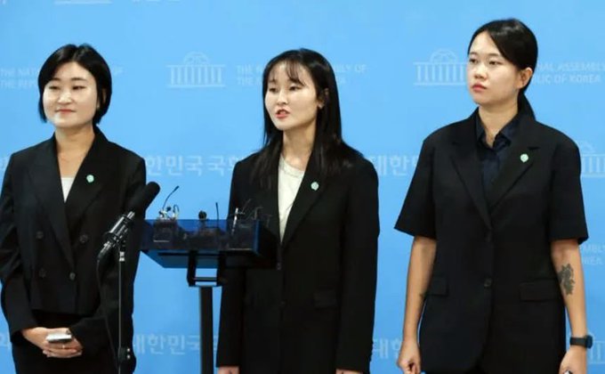 Three women in dark suits stand at a press briefing with the National Assembly of the Republic of Korea emblem behind them, with the spokesperson speaking into a microphone. The captioner's comments about tattoos and short-sleeve jackets appear to be jokingly misidentifying gender, as all three individuals pictured are women—and a tattoo is visible on the right arm of the woman on the far right.