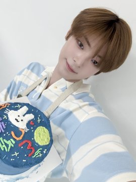 A young boy with light brown hair smiles softly while wearing a blue and white striped shirt, holding what appears to be a birthday cake decorated with an astronaut design. The image ties to the post about a cake prepared for Kang Woojin (강우진), likely celebrating his birthday as part of a fan update.