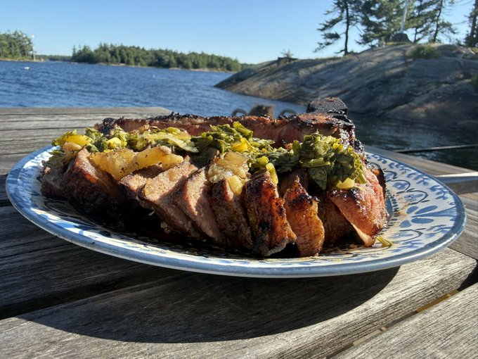 A sliced grilled steak topped with a chunky green herb and yellow pepper sauce, served on a blue and white patterned plate on a wooden dock by a lake.