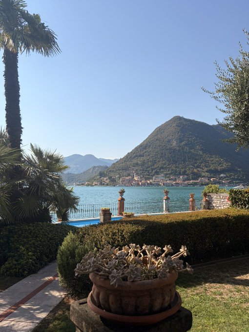 A serene lakeside view of Lake Como in Italy, framed by manicured gardens, classical urns, and a towering mountain in the distance. The tranquil setting mirrors the calm-before-the-storm optimism in the post, as the peaceful "GM" scene reflects the trader's confident belief that the market bottom is in and a new wealth-building cycle is about to begin.