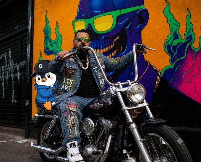 A tattooed man in a denim vest and sunglasses sits on a motorcycle in front of a vibrant mural featuring a blue skull wearing green sunglasses, with a cartoon penguin character added to the scene.