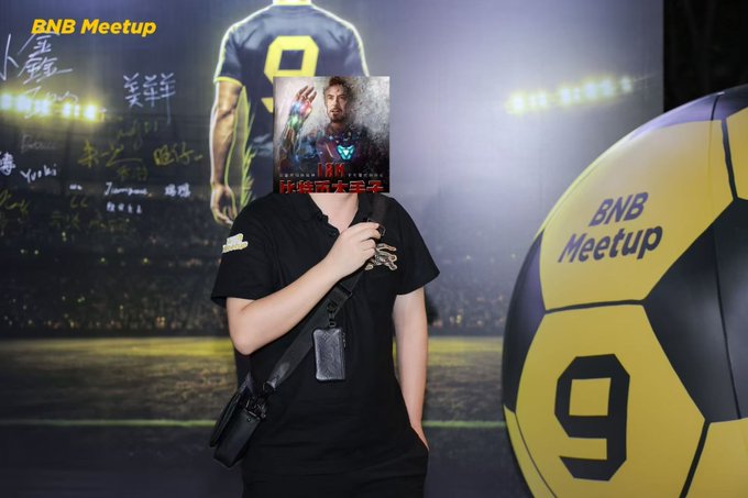A person in a black t-shirt stands in front of a "BNB Meetup" backdrop featuring a soccer player wearing jersey number 9 and a large soccer ball, holding an Iron Man-themed item that obscures their face.