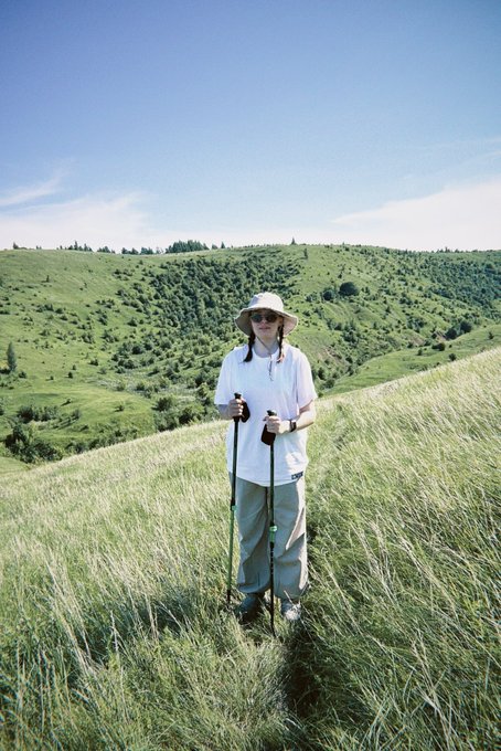 A person wearing a white t-shirt, gray pants, and a sun hat stands in a grassy mountain meadow holding two hiking poles, with rolling green hills and blue sky in the background.