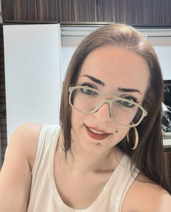 A close-up portrait of a woman with reddish-brown hair, wearing a cream-colored tank top and distinctive green-rimmed glasses with gold hoop earrings.