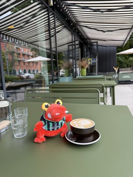 A red plush toy wearing a blue and white striped shirt sits on a green table next to a cup of coffee and a glass of water at an outdoor café.