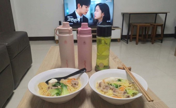 Two bowls of noodle soup with beef, bok choy, and egg sit on a wooden table alongside two pink Thermos bottles and a bottle of green tea, with a Korean drama playing on the TV in the background.