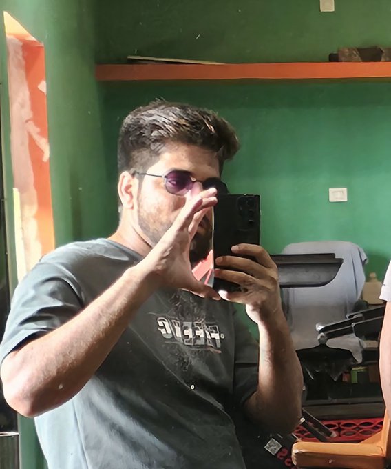 A mirror selfie of a man wearing purple sunglasses and a dark gray t-shirt, holding a black smartphone, taking a photo in a room with green walls.