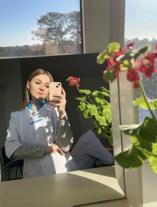 A young woman in a white lab coat takes a mirror selfie with her phone, posing beside a flowering geranium plant in a sunlit room. The image reflects her early days studying her chosen field before pivoting to a career in crypto.
