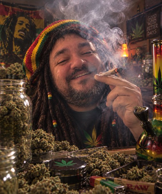 A man with dreadlocks wearing a red, yellow, and green beanie smiles with his eyes closed as he smokes a large joint, surrounded by jars of cannabis and Bob Marley imagery.