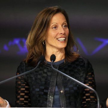A smiling woman with brown hair, wearing a patterned black jacket, speaks at a podium with microphones, consistent with her role as Nasdaq CEO Adena Friedman discussing tokenization of traditional assets like Treasurys and stocks onto blockchain networks.