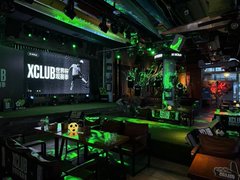 Interior of a dimly lit themed bar or restaurant with green ambient lighting, leather seating, multiple TVs, and an Xbox gaming area visible.