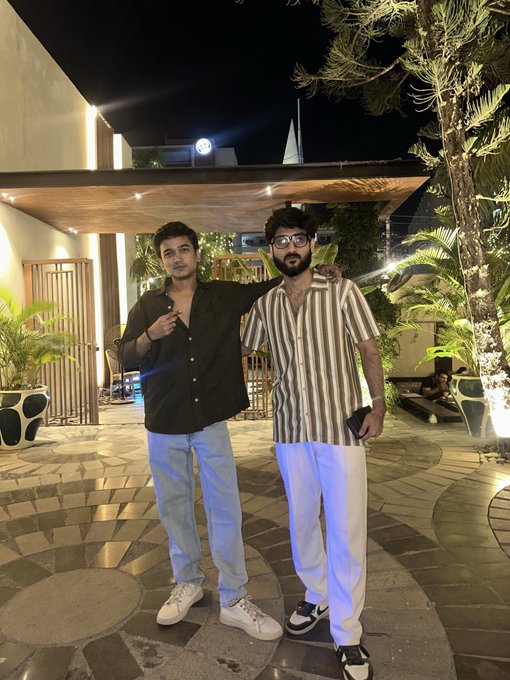 Two young men pose together at night on a patterned patio outside a building, with one wearing a black shirt and the other in a striped shirt and white pants.