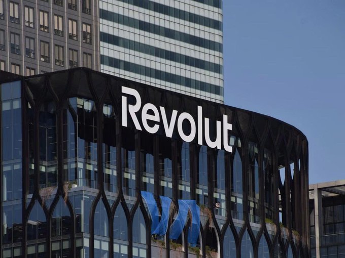 A modern glass office building displays the 'Revolut' logo prominently on its dark facade against a clear blue sky.