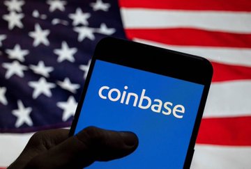 A hand holds a smartphone displaying the Coinbase logo on a blue screen with an American flag in the background.