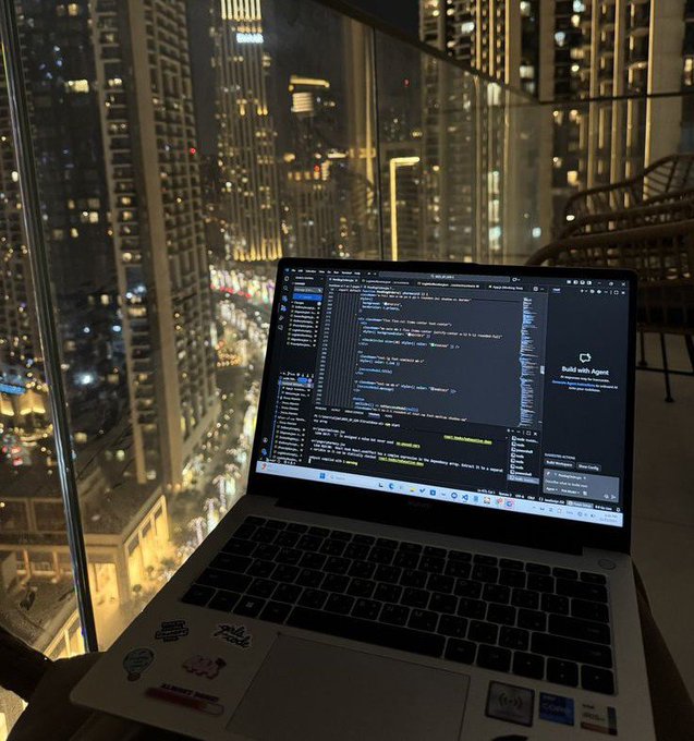 A laptop displaying code is open on a high-rise balcony overlooking a brightly lit city skyline at night, with stickers visible on the laptop's palm rest.