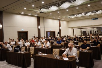 Attendees are seated at long tables covered with brown cloths inside a large conference hall, attentively participating in the WinC300 community event in Ankara, where presentations on portfolio management, chart analysis, and bots were held.