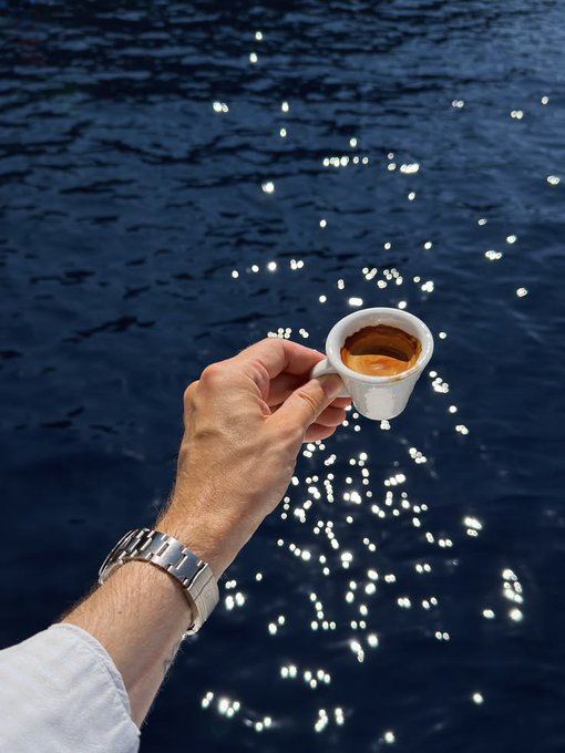 A person's arm, wearing a silver watch and white sleeve, extends over dark blue water holding a small white espresso cup filled with coffee, with sunlight reflecting on the water's surface.