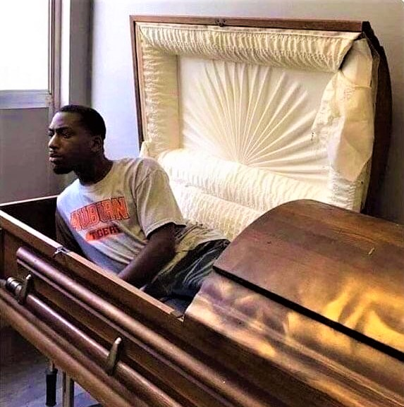 A humorous photoshopped image showing a young man wearing an 'Auburn Tigers' t-shirt sitting inside an open wooden casket/coffin, with the interior lid displaying white satin lining.