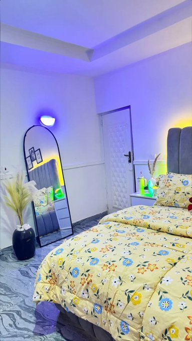 A modern bedroom with a floral yellow bedspread, an arched mirror with LED lighting, and purple and yellow ambient lighting throughout the room.