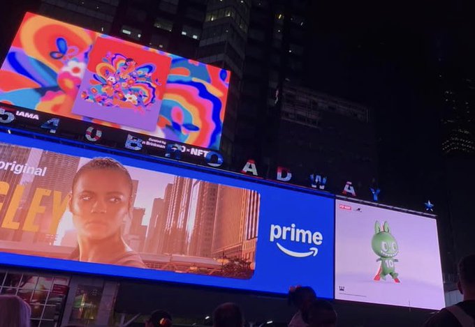 Nighttime photograph of Times Square billboards displaying colorful psychedelic art, an Amazon Prime ad, and what appears to be the Smurfs movie promotion.