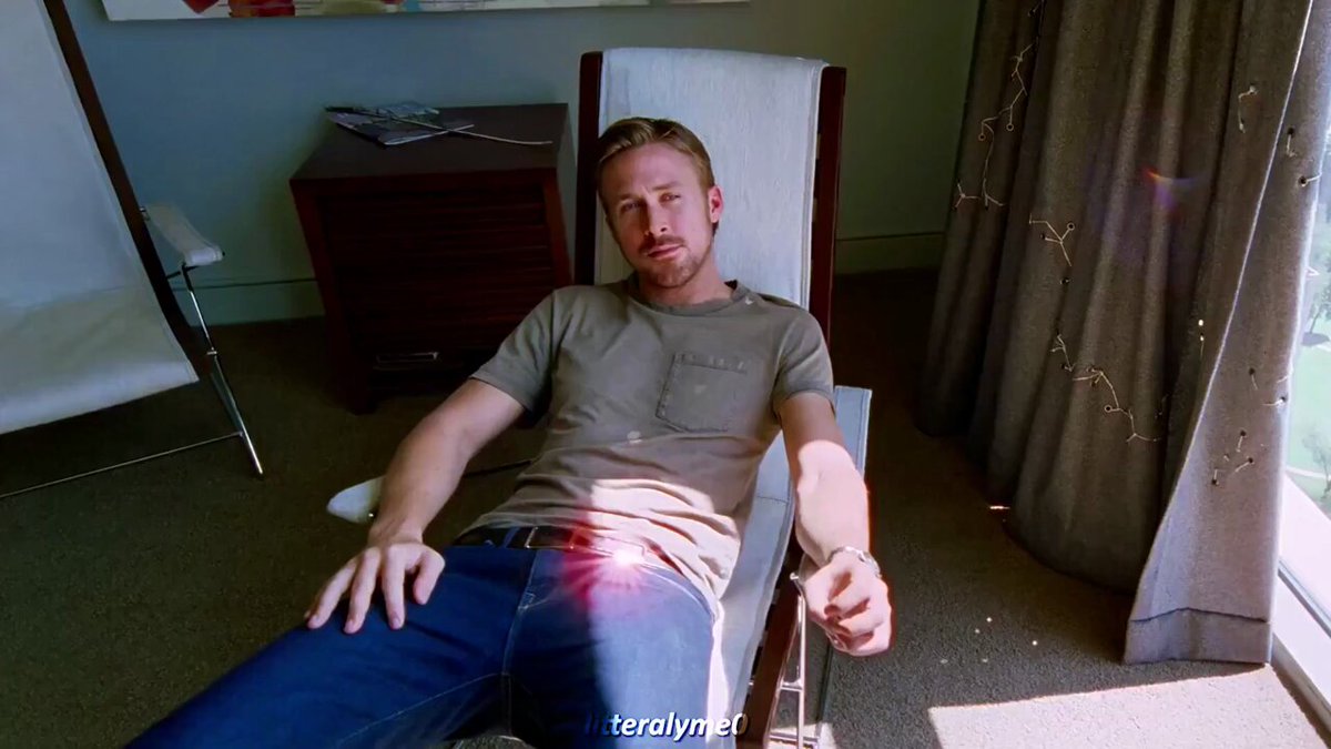 The image shows a man sitting in a chair with his arms crossed, wearing a gray shirt and blue jeans. He has his hands resting on his knees while looking directly at the camera. The background features a window that allows natural light into the room, creating an atmosphere of relaxation or contemplation. There is also a book visible in the scene, suggesting that the man might be reading or enjoying some leisure time.
