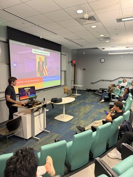 A photograph of a university lecture hall where a presenter stands at a podium with a laptop, showing a presentation slide to seated students in green chairs.