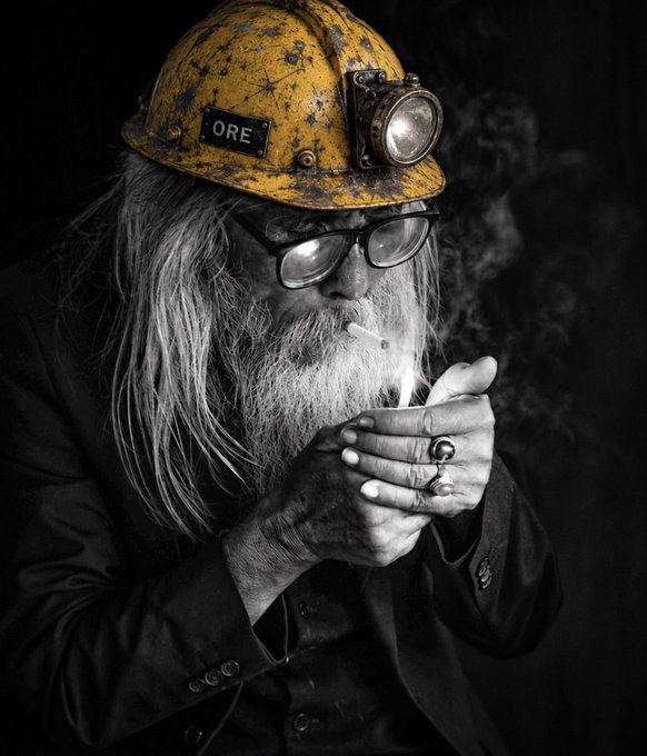 A black and white photograph of a bearded man with long hair wearing a yellow mining helmet with 'ORE' written on it, lighting a cigarette against a dark background.