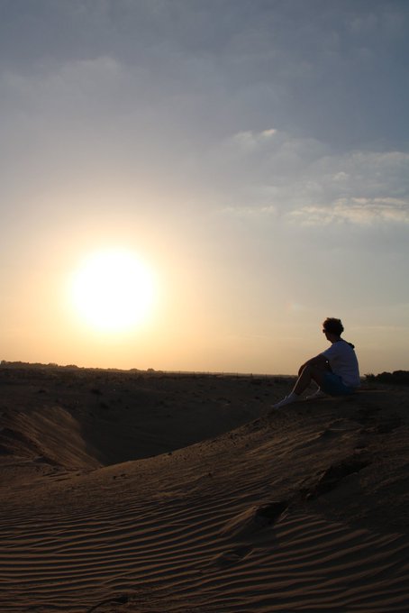 A solitary figure sits atop a sand dune, silhouetted against a hazy desert sunset, capturing a moment of quiet contemplation in an expansive, arid landscape. The image aligns with the "GN" post, evoking themes of solitude, natural beauty, and reflective introspection in a remote setting.