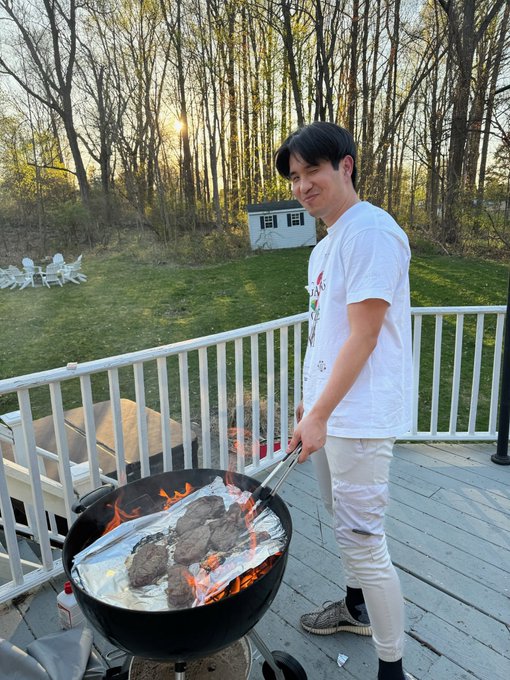 A man wearing a white t-shirt stands outside, cooking food on an outdoor grill with a large metal pot of meat. The photo also includes a caption that reads "GM #AVAX I'm enjoying the warm weather today." This suggests that the man is enjoying his time outdoors and appreciates the pleasant weather while preparing his meal.
 
The image captures a moment in which the man is cooking food on an outdoor grill, surrounded by trees and possibly other people nearby. The scene conveys a sense of relaxation and enjoyment as he takes advantage of the warm weather to prepare a delicious meal.