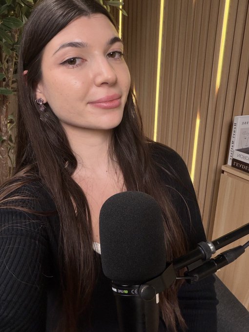 A young woman with long dark hair sits in front of a Shure microphone in a room with wood-paneled walls, appearing to record a podcast or video.