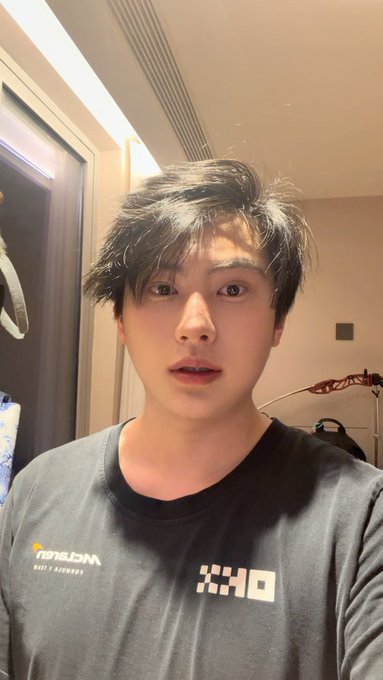 A young man wearing a grey long-sleeved shirt with a 'DRIX' logo takes a selfie indoors. He has messy dark hair and is looking directly at the camera.