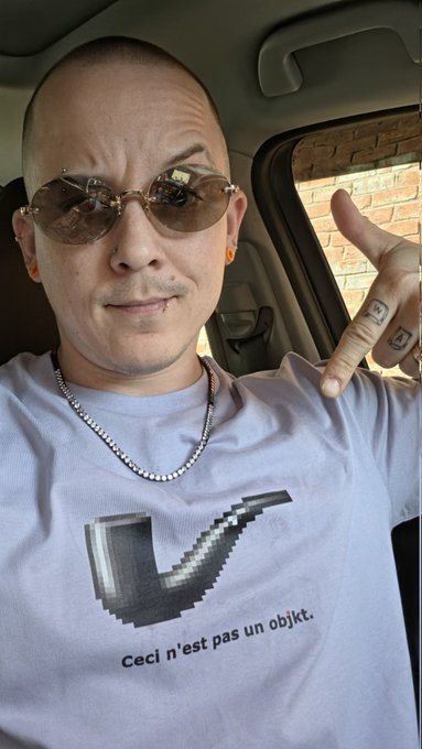 A selfie of a man with short hair wearing brown tinted sunglasses, a silver chain necklace, and a white t-shirt featuring a pixelated pipe graphic with the French text 'Ceci n'est pas un objet,' taken inside a car.