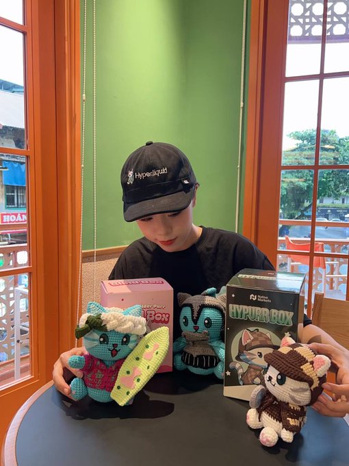 A person wearing a black 'Hyperliquid' cap and black t-shirt sitting at a table, displaying merchandise. They are holding or have in front of them crocheted amigurumi figures, a pink 'Magic Box' box, and a black 'Hyper Box' packaging featuring cartoon characters.