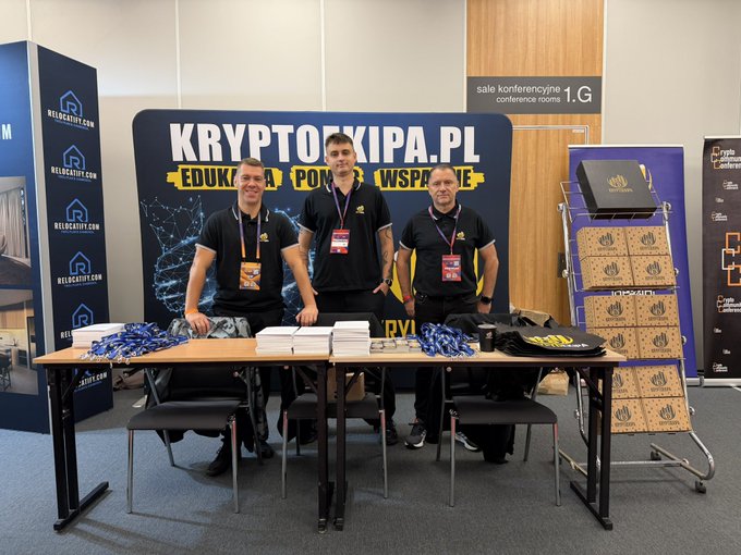 Three individuals stand behind a table at a booth for 'KRYPTOKIPA.PL' promoting education, business, and support.