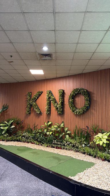 An indoor corporate display featuring the letters 'KNO' decorated with greenery mounted on a wood-paneled wall, with potted plants and a strip of artificial grass below.