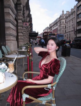 A young woman in an elegant dark red velvet dress sits at an outdoor Parisian café, posing gracefully with one hand near her neck. The accompanying caption "Call Mommy" adds a playful, humorous tone to the sophisticated street-side scene.