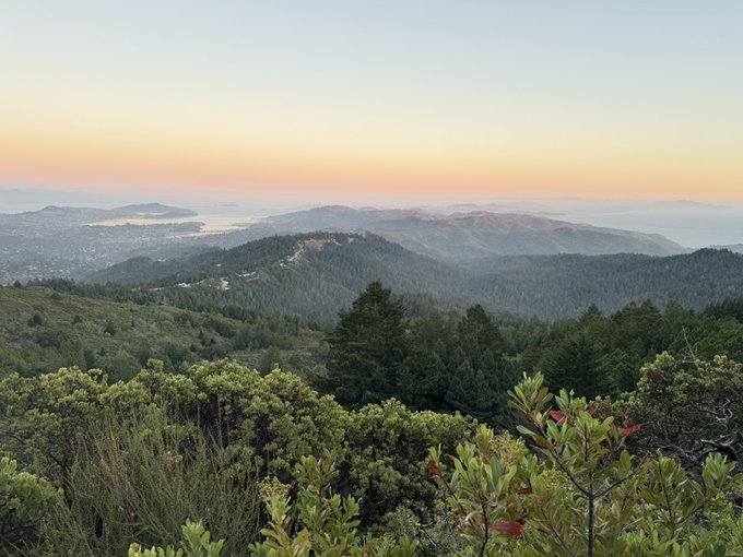 A panoramic vista from Mount Tamalpais at sunset captures rolling forested hills descending to the Pacific Ocean and bay waters, with a city nestled along the coast between them.