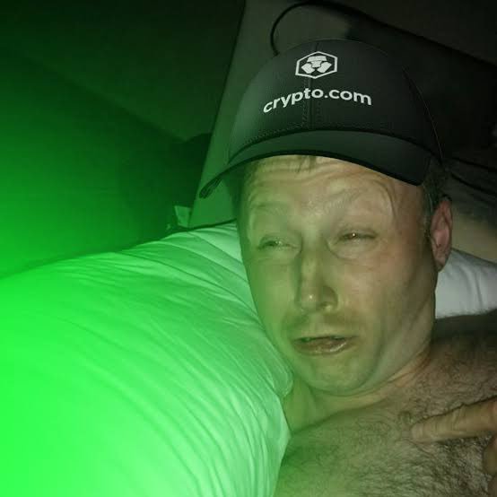 A man wearing a black "crypto.com" cap lies in bed bathed in green light, appearing tired or unwell. The caption "gM CT We're so back" combined with the green hue and Crypto.com branding humorously nods to the return of crypto-related content or sentiment in a casual morning greeting.