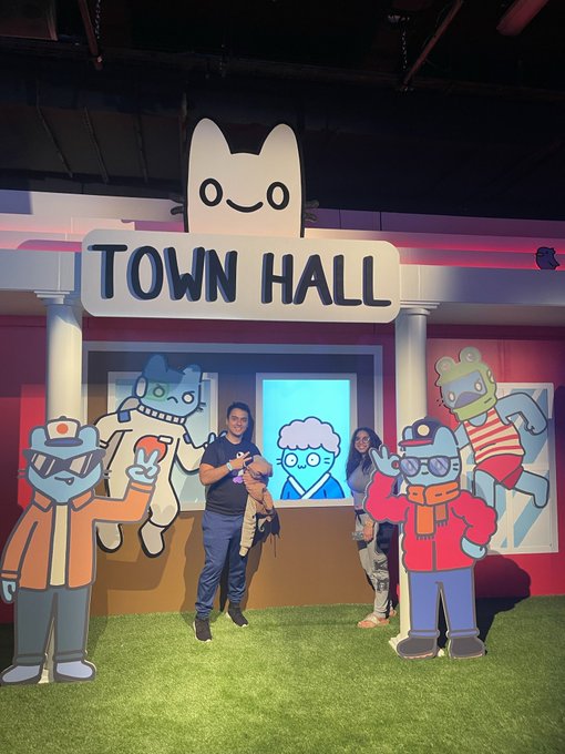 Two people pose in front of a colorful 'TOWN HALL' display featuring cartoon character cutouts at what appears to be a themed entertainment venue.