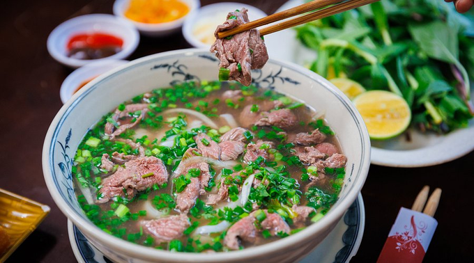 A bowl of Vietnamese pho filled with beef slices, onions, and cilantro in a savory broth, with chopsticks lifting a piece of meat, accompanied by fresh herbs, lime, and chili sauce on the side. The post appears to be a casual "gm ct" greeting inviting discussion about pho, with the steaming bowl serving as a fitting visual prompt for the conversation.